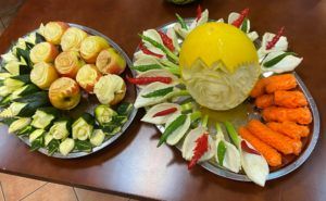 Po lewej stronie, na talerzu kompozycja kwiatowa wyrzeźbiona w jabłkach i cukinii. Po prawej  stronie, na talerzu kompozycja kwiatowa wyrzeźbiona w melonie i w porach oraz szyszki z marchwi.